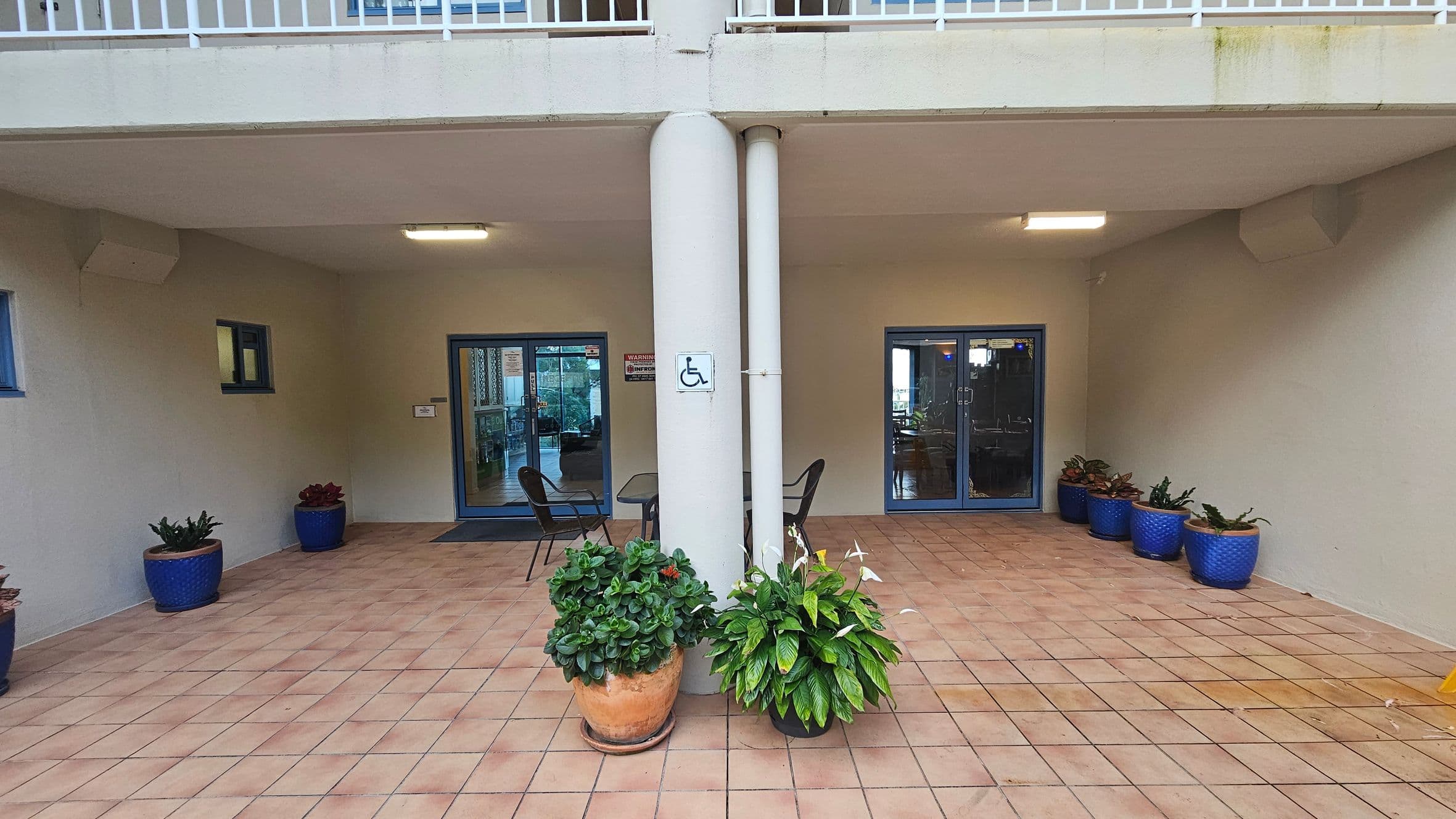 A covered outdoor area with a tiled floor and a beige ceiling. In the centre, two large columns with a wheelchair accessibility sign. Between the columns are two potted plants. On either side, there are blue-framed glass doors leading inside. Blue plant pots with greenery are placed along the walls. Two chairs and a small table are positioned near the left door. A railing is visible above the area.