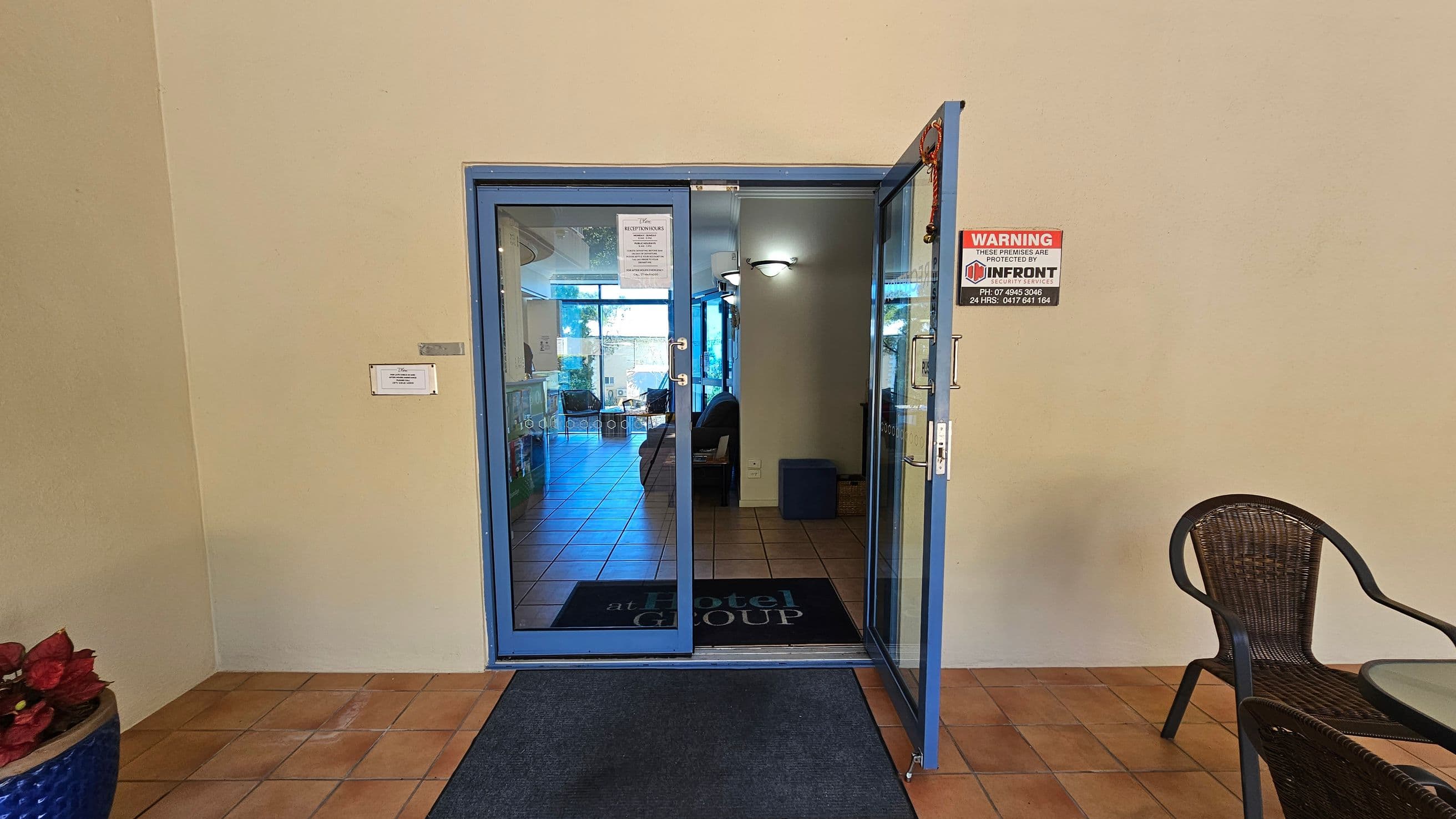 A glass door with a blue frame is slightly open, leading into a room with tiled flooring. On the left, a blue pot with red plants sits on a terracotta-tiled floor. A sign is visible on the wall next to the door. On the right, a wicker chair is partially visible. Inside, a mat with "at Hotel Group" is on the floor, and a seating area is visible in the background. A security warning sign is on the door.