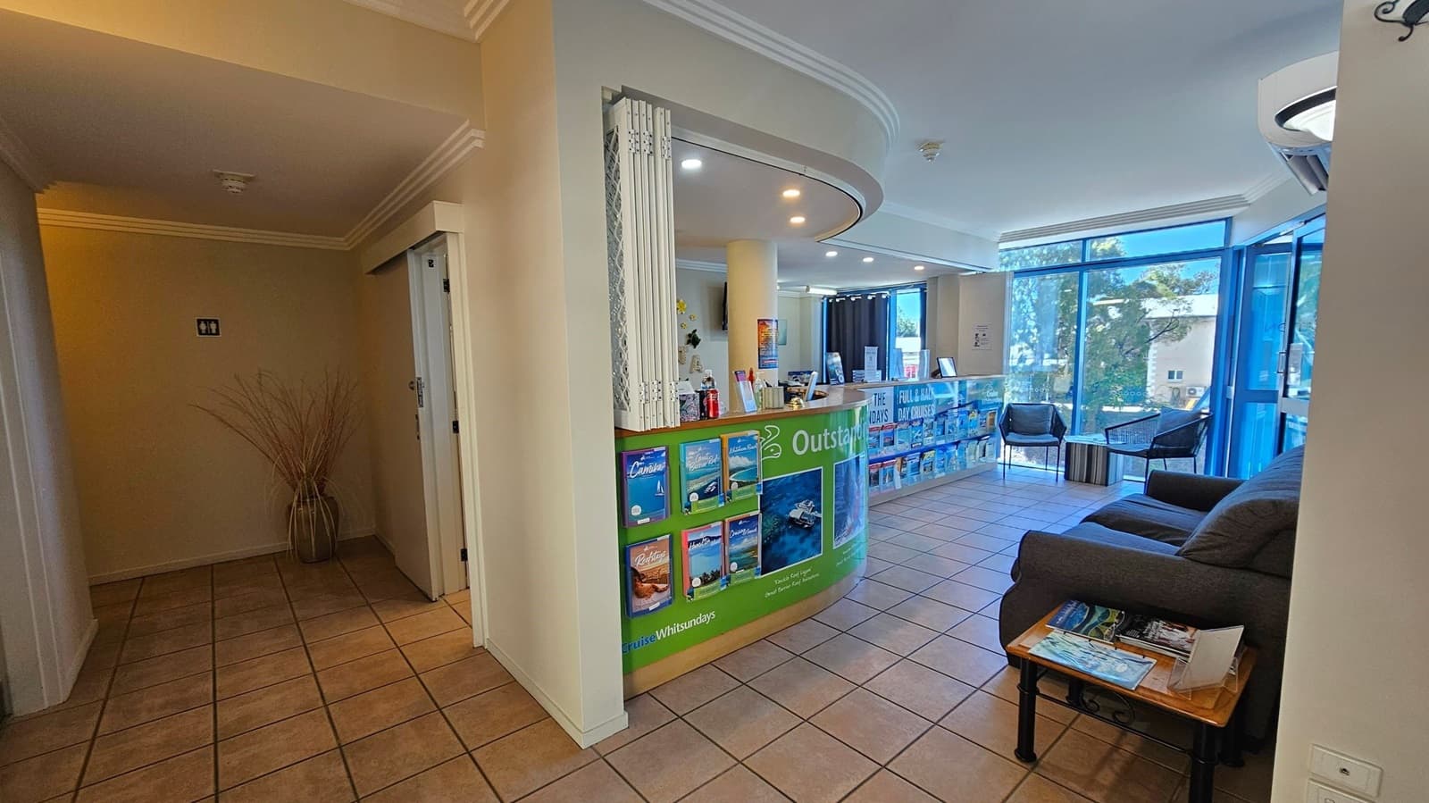 A reception area with a tiled floor and a curved counter displaying brochures. On the right, there's a dark sofa with a small table holding magazines. Large windows with blue frames let in natural light, and two chairs are positioned near the windows. The ceiling has recessed lighting, and a vase with tall branches is in the corner on the left.