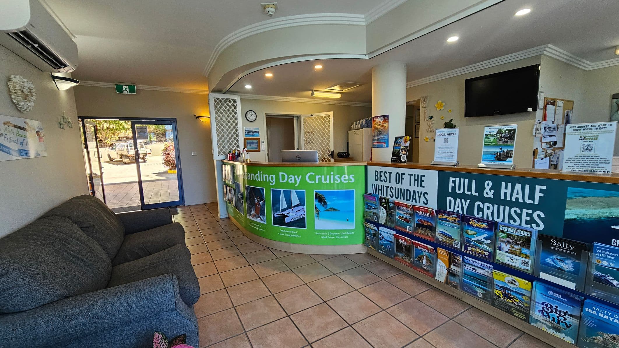 Reception area with a curved desk in the centre displaying brochures for day cruises. Behind the desk, a computer and a clock are visible. To the right, a wall-mounted TV and more brochures. On the left, a grey sofa sits against the wall near a glass door leading outside. The floor is tiled, and the ceiling has recessed lighting.