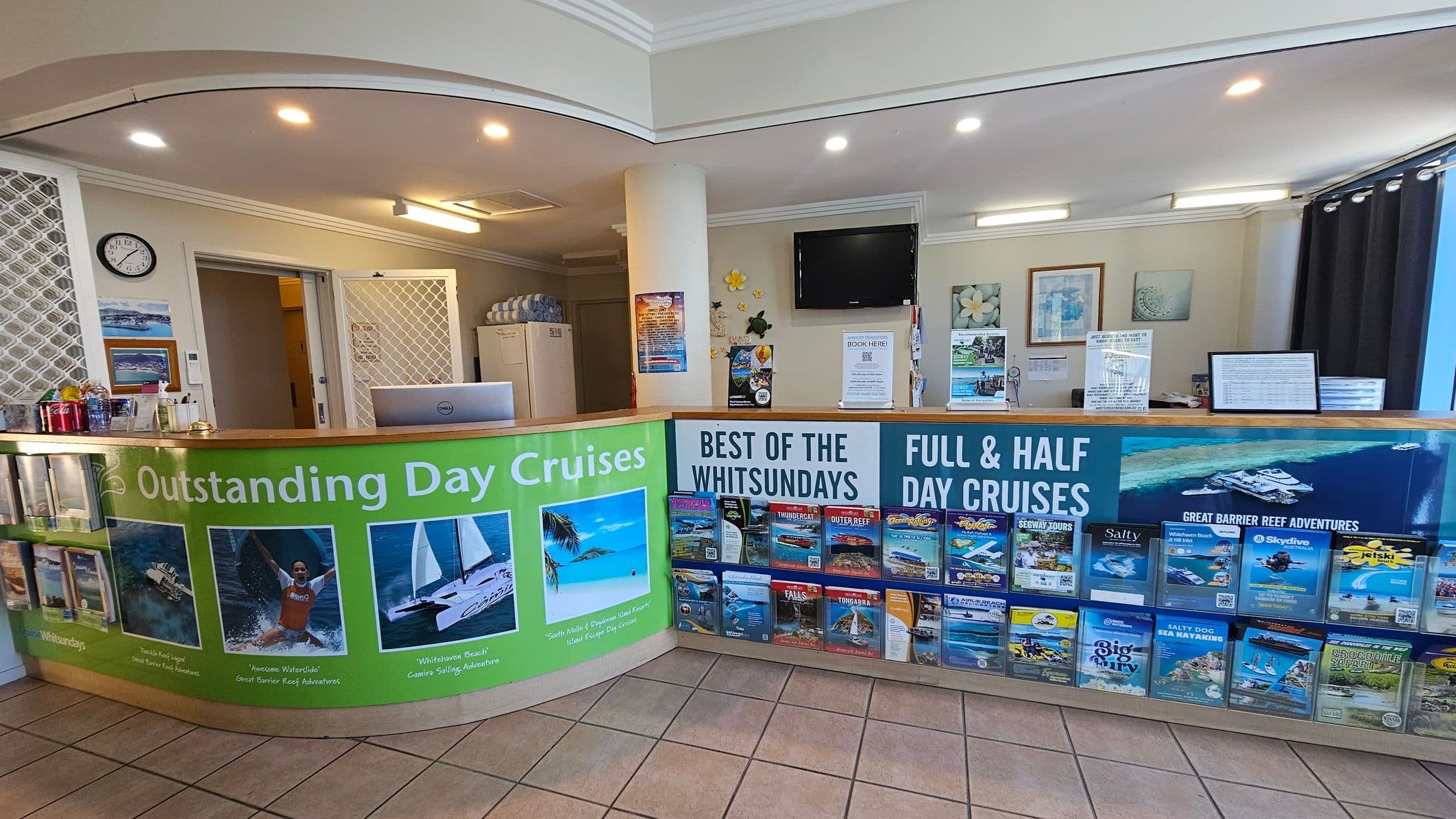 Reception area with a curved counter displaying brochures and advertisements for day cruises. The counter is decorated with vibrant images of boats and water activities. Behind the counter, a wall-mounted TV and various posters are visible. To the left, a small fridge with drinks and a clock on the wall. The floor is tiled, and the ceiling has recessed lighting. A doorway leads to another room, and there are some towels stacked near the entrance.
