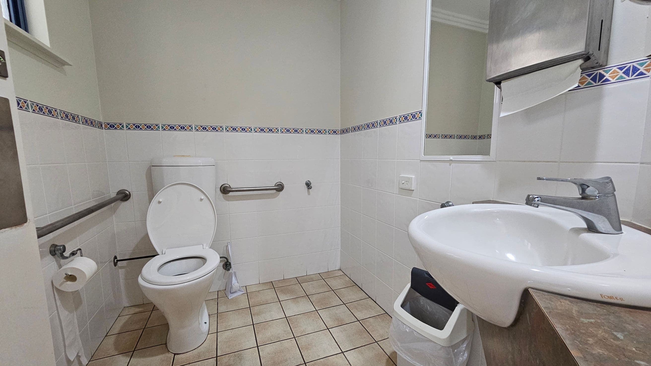 A bathroom with a white toilet on the left, equipped with metal grab bars and a toilet roll holder. The walls are tiled with a decorative border. On the right, a white sink with a silver tap is mounted on a wooden counter. Above the sink is a mirror and a metal paper towel dispenser. The floor is covered with beige tiles.
