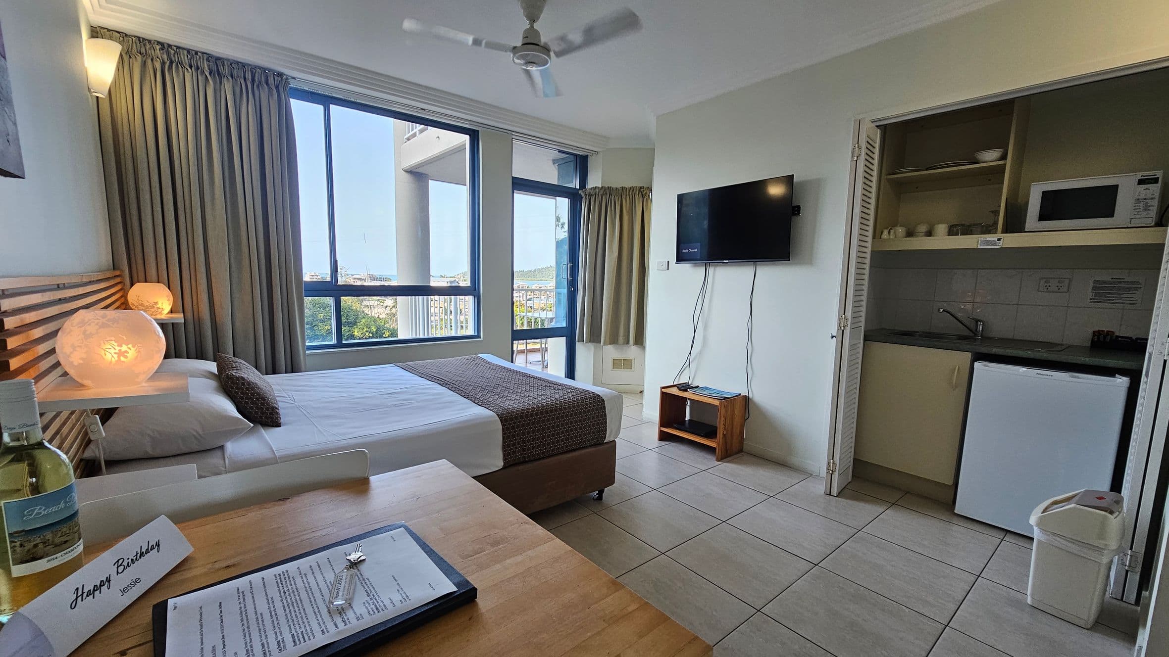 A hotel room with a tiled floor and a ceiling fan. On the left, there's a wooden table with a white chair, a bottle, and a card. Above the table, a colourful abstract painting hangs on the wall. In the centre, a bed with a wooden slatted headboard and two pillows is positioned against the wall. A large window with curtains and a glass door leads to a balcony. On the right, a flat-screen TV is mounted on the wall above a small wooden shelf. A louvred door is partially open.