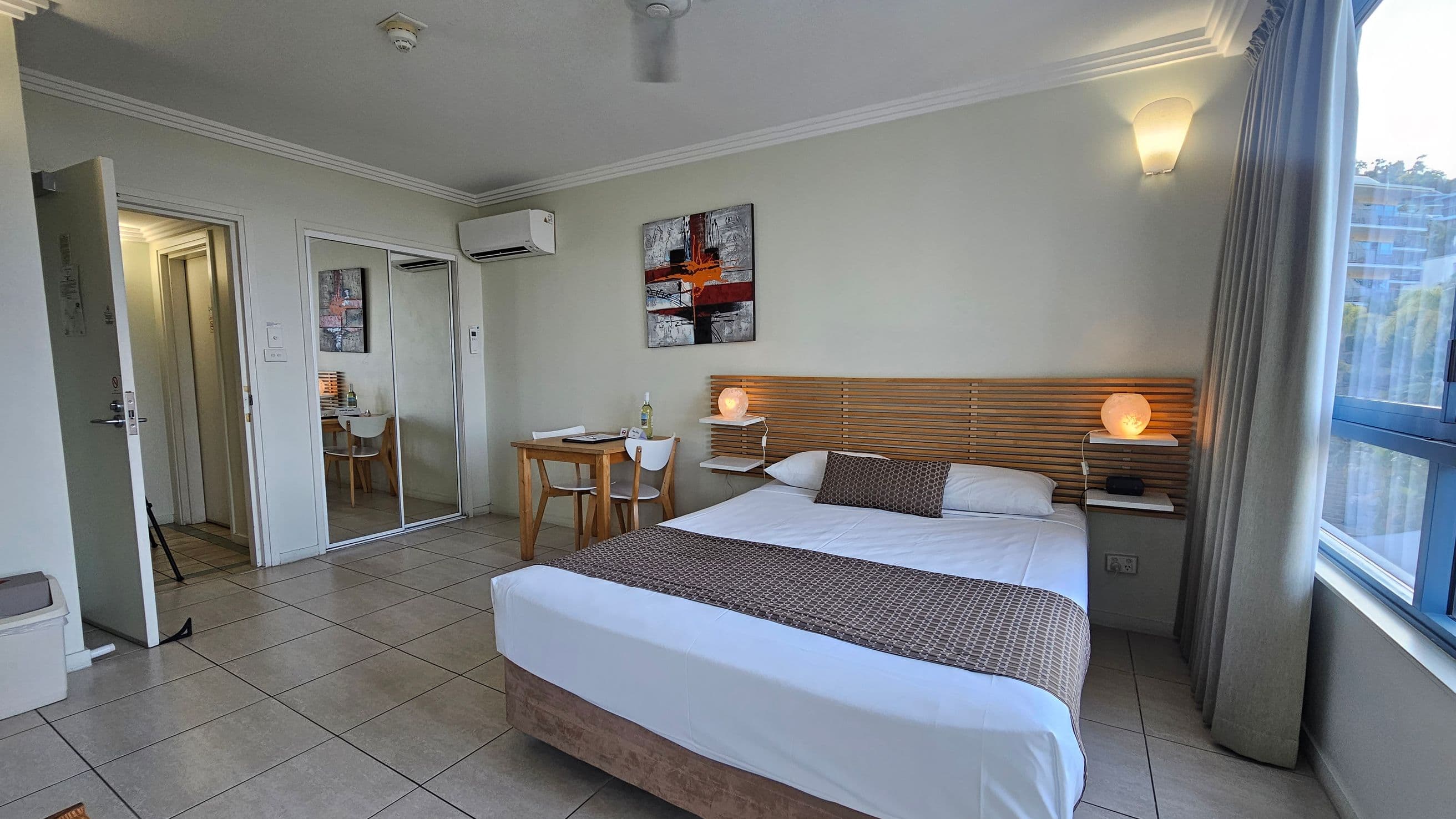A tidy room with a bed on the right, featuring a wooden headboard and a small lamp on a shelf. A window with curtains is beside the bed. On the left, there's a small wooden table with a chair, and a bottle on the table. A painting hangs above the table. The floor is tiled, and there's an open door leading to a hallway. A mirrored wardrobe is next to the door, and an air conditioning unit is mounted above it. The ceiling is plain with a light fixture.