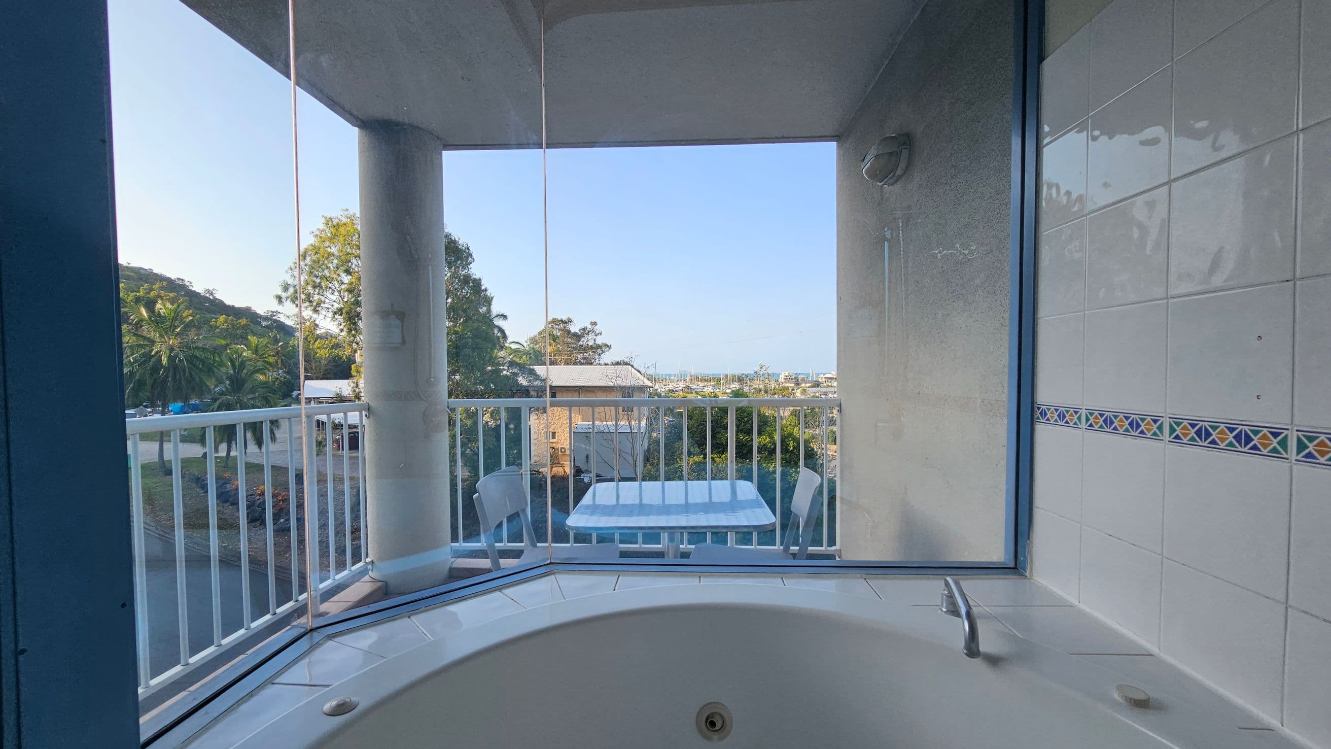 A bathroom with a large corner window overlooking a balcony. The foreground features a white bathtub with a silver tap. The walls are tiled with a decorative border. Outside, a small table with two chairs is visible on the balcony. Beyond the railing, there are trees and a distant view of buildings and the sea. A large pillar supports the balcony roof.