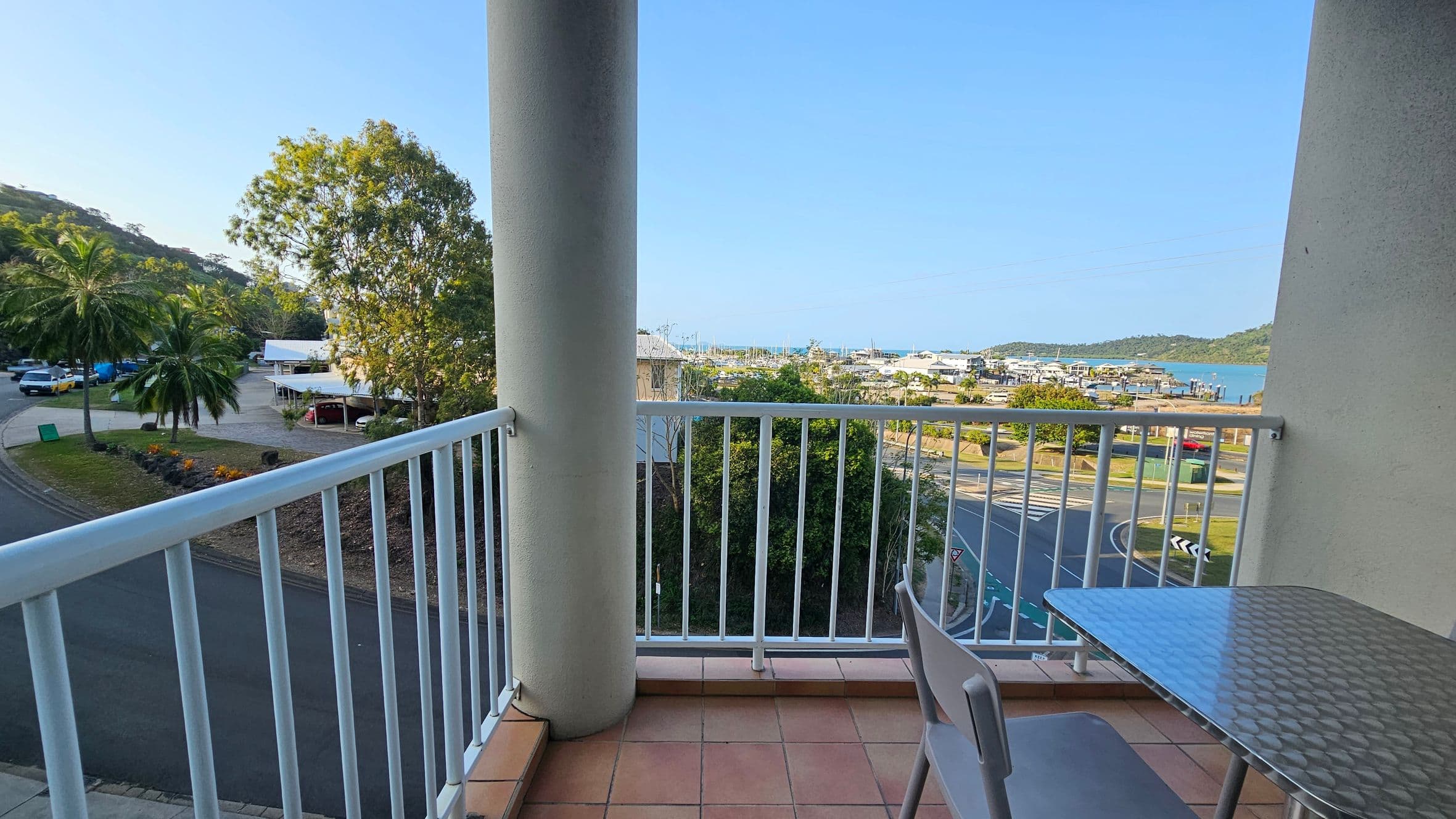A balcony with a tiled floor and a white railing overlooks a street and a marina. On the right, there's a metal table with a chair. In the background, palm trees and other greenery are visible, along with a few buildings and parked cars. The marina is seen in the distance, with boats and a view of the water. A large tree is on the left, and a roundabout is visible below.