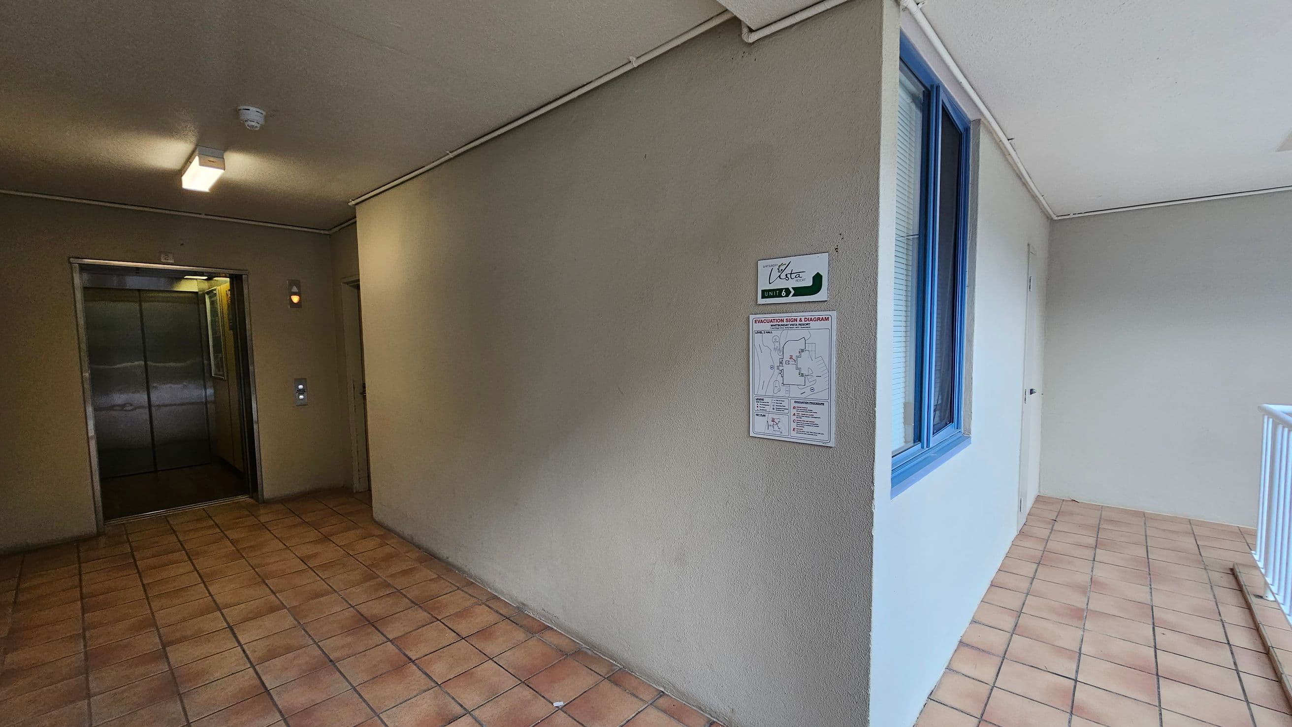 On the left, there is an open lift with metallic doors and a control panel beside it. The floor is covered with brown tiles. In the middle, a beige wall displays a sign labelled "Vista Unit 3" and an evacuation diagram. On the right, a blue-framed window is set into the wall, and a white railing is visible. The ceiling is plain with a light fixture.