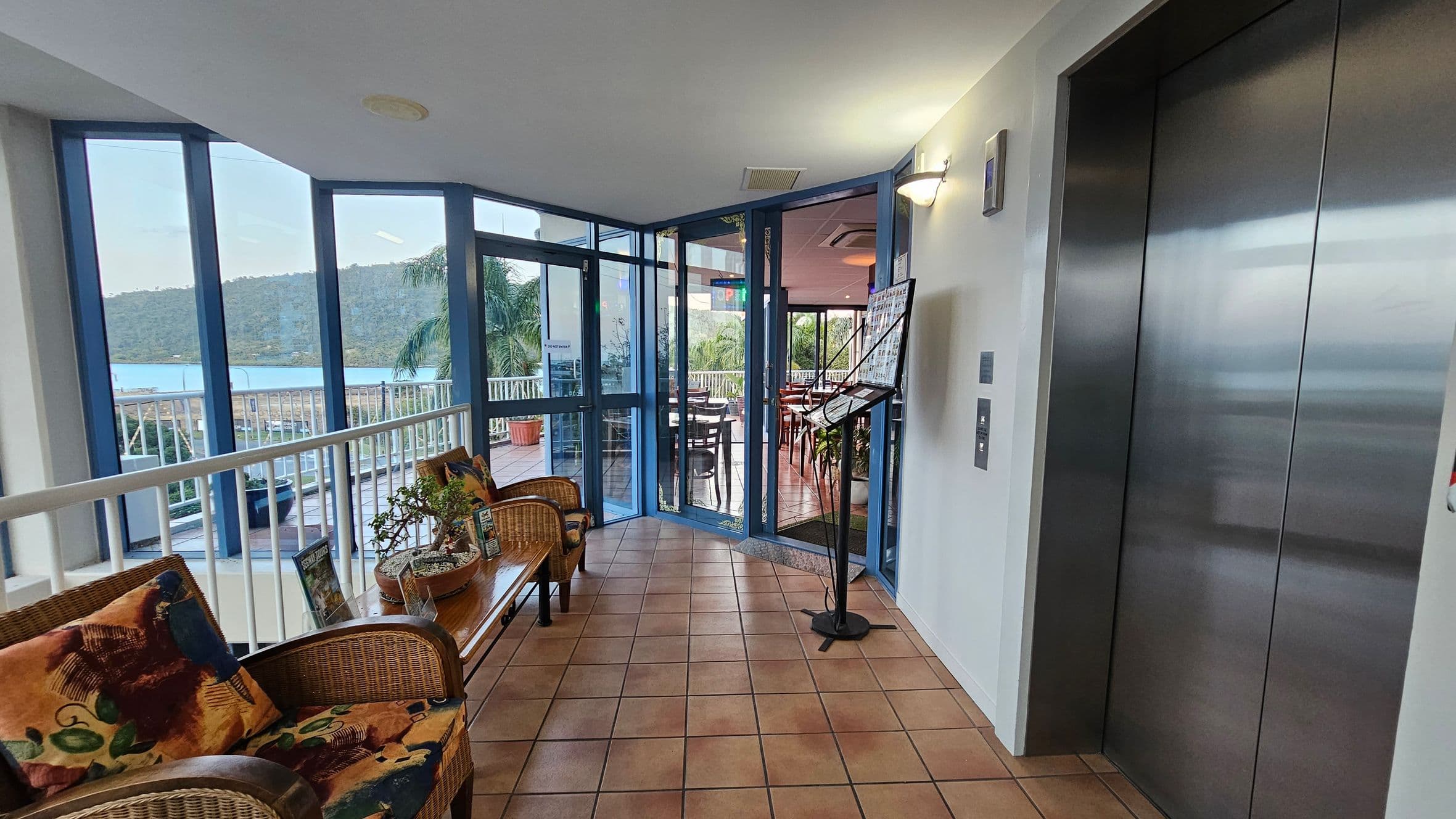 A hallway with tiled flooring and a ceiling light. On the right, there is a closed lift with metallic doors. On the left, a seating area with wicker chairs and colourful cushions, accompanied by potted plants. Large windows offer a view of a balcony with railings, potted plants, and a distant hillside. In the centre, a glass door leads to a dining area with tables and chairs, and a stand with brochures is positioned nearby.