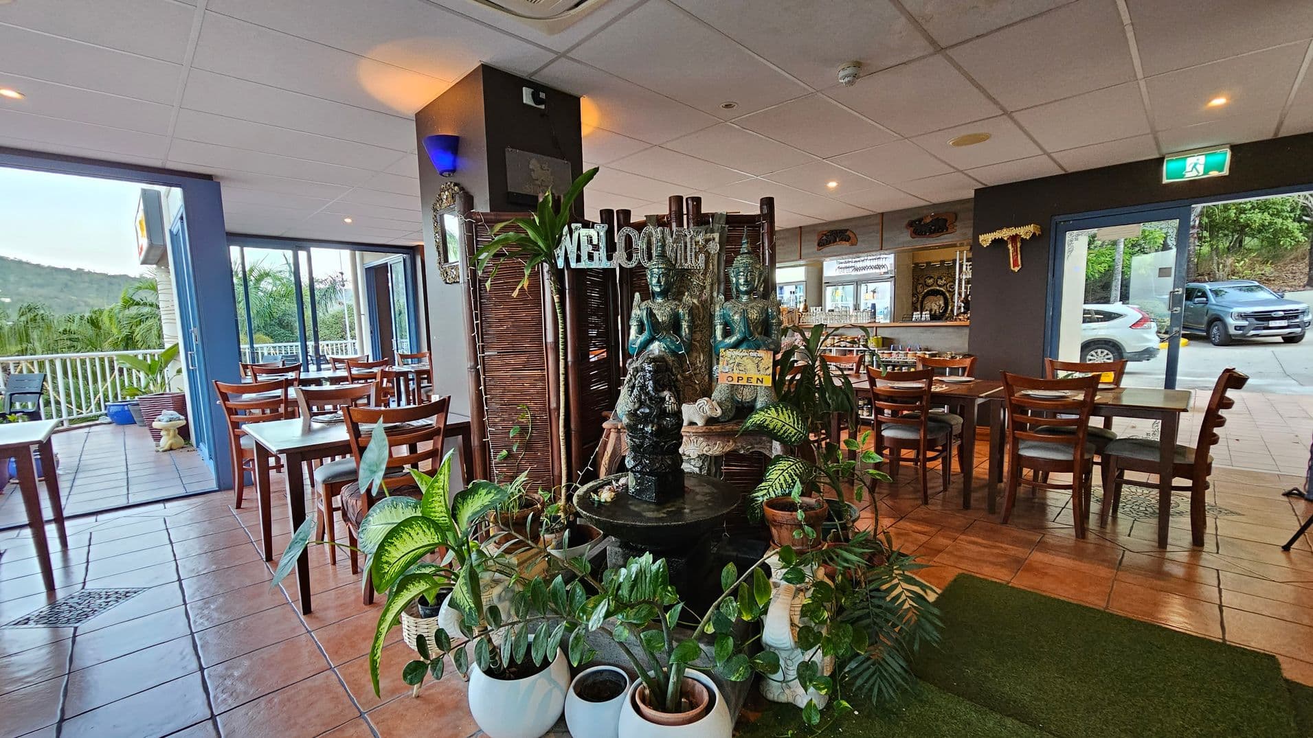 Indoor restaurant with a central decorative area featuring a small fountain surrounded by potted plants and statues. Wooden tables and chairs are arranged on a tiled floor. To the left, large windows and doors open to a balcony with a view of greenery. On the right, a bar area is visible, and a glass door leads outside where cars are parked. The ceiling is white with recessed lighting.