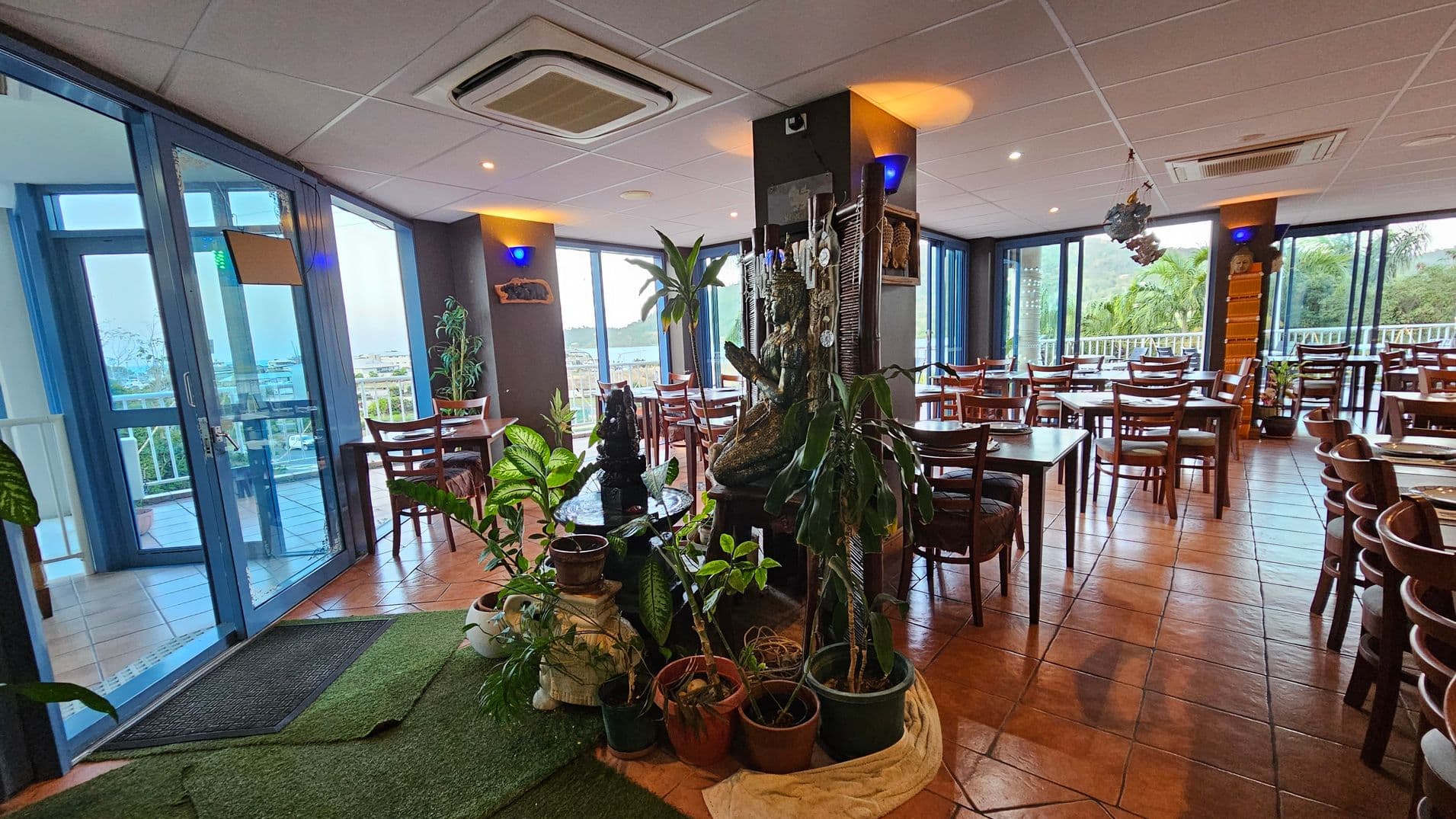 A restaurant interior with terracotta tiled flooring and a white panelled ceiling. On the left, large blue-framed glass doors lead to an outdoor area. In the centre, a collection of potted plants surrounds a decorative statue. Wooden tables and chairs are arranged throughout the space, with large windows on the right offering views of greenery outside. Blue wall lights and air conditioning units are visible on the ceiling.