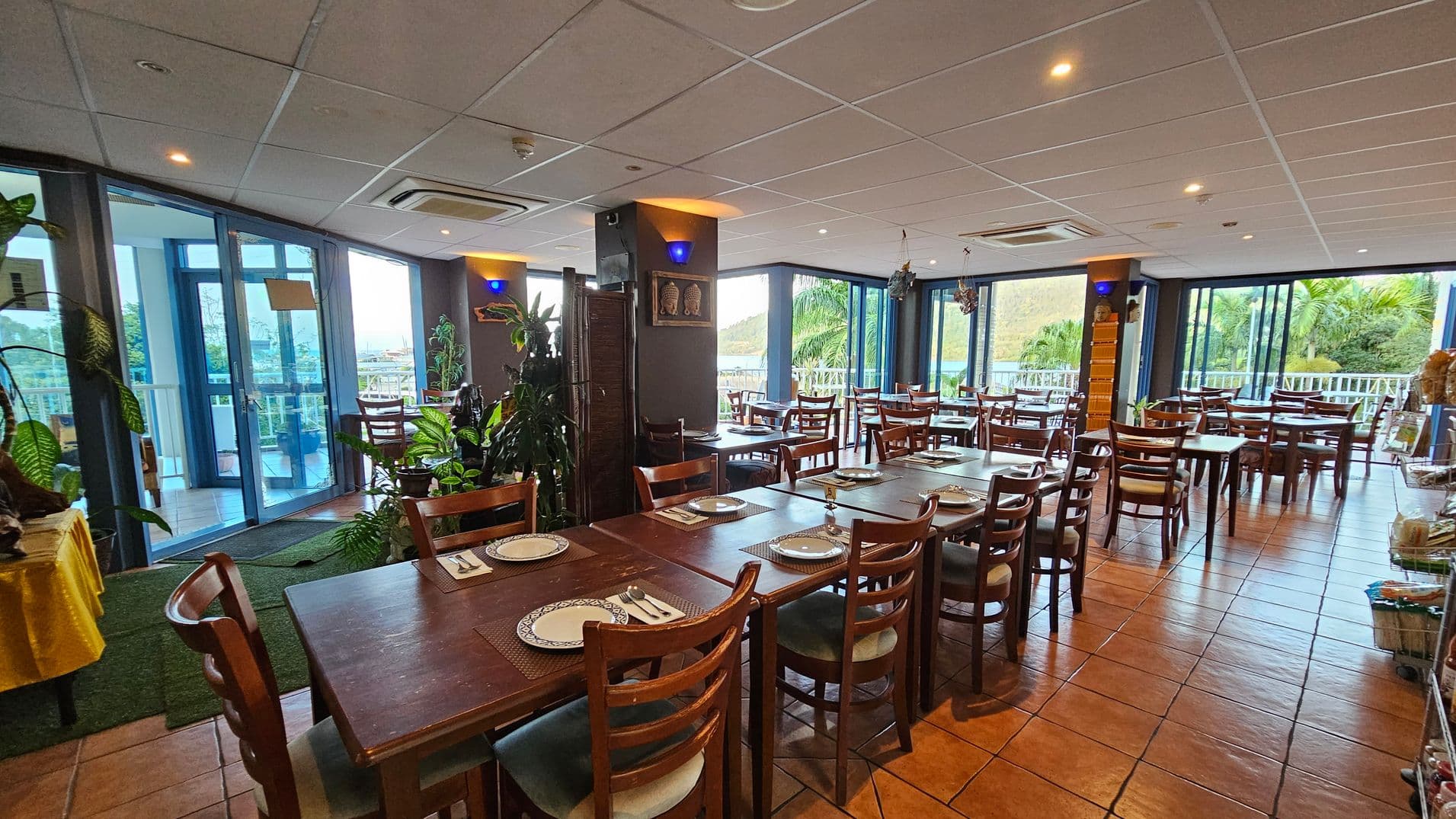 A spacious restaurant interior with wooden tables and chairs arranged neatly. Each table is set with plates and cutlery. Large windows on the right and in the background offer views of greenery and let in natural light. Potted plants are scattered throughout, adding a touch of nature. The floor is tiled in a warm terracotta colour, and the ceiling is white with recessed lighting. A yellow tablecloth covers a table on the left, and decorative items adorn the walls.