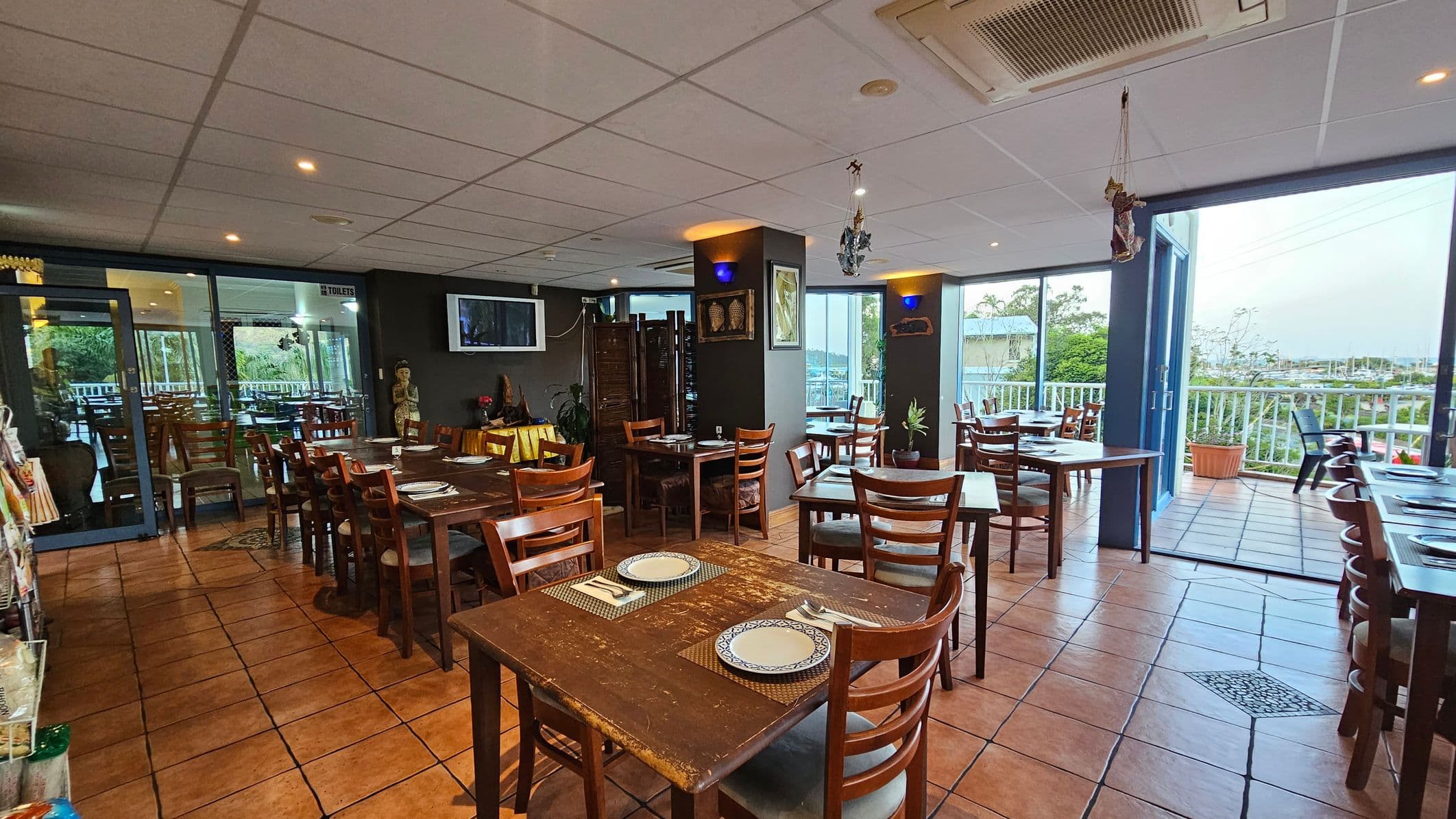 A restaurant interior with wooden tables and chairs arranged neatly. On the left, a glass door leads to another dining area. In the centre, a decorative statue and wall art are visible. The right side features large windows and a balcony with outdoor seating, offering a view of greenery. The floor is tiled in a terracotta colour, and the ceiling is white with recessed lighting.