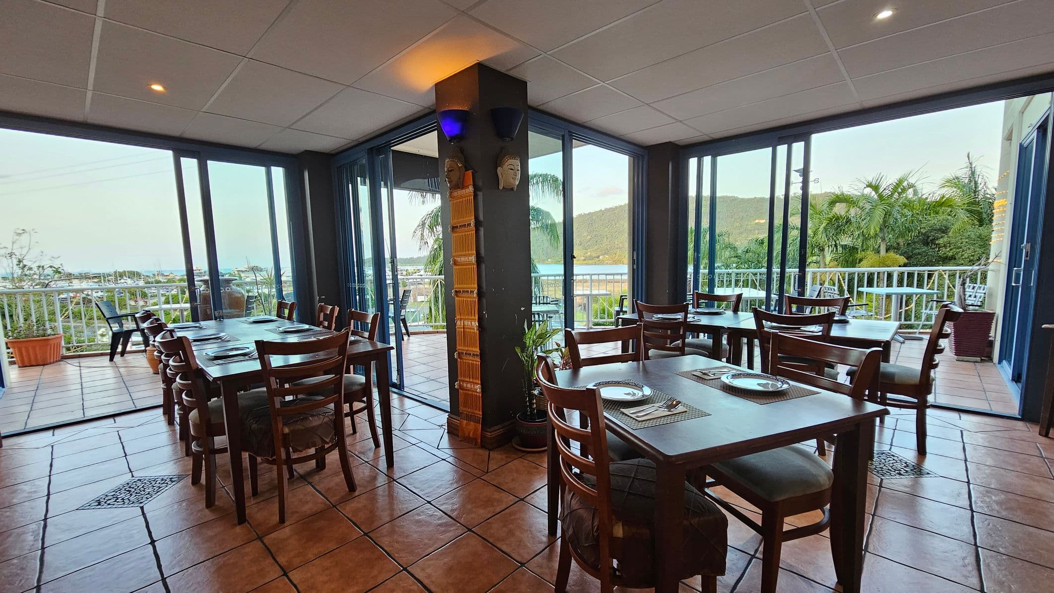 Dining area with wooden tables and chairs set on a tiled floor. Large glass doors on the left and right open to a balcony with views of greenery and distant hills. A central pillar features decorative masks and a small plant at its base. The ceiling is white with recessed lighting.