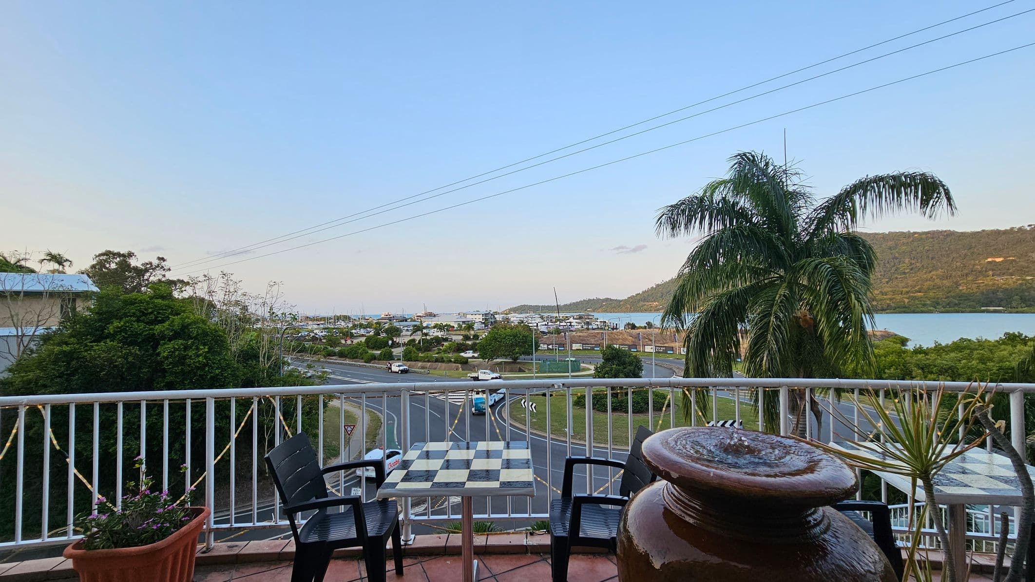A balcony view with a small table and two chairs in the foreground. The table has a black and white checkered pattern. To the right, a large ceramic pot with a plant is visible. In the background, a road with a roundabout is seen, along with several cars. Beyond the road, there are buildings, a marina with boats, and a body of water. A large palm tree is on the right, and hills are visible in the distance. The balcony railing is adorned with string lights.
