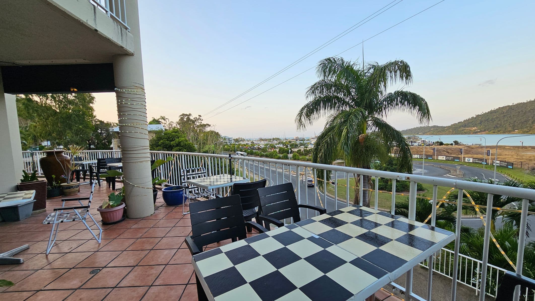 A balcony with a tiled floor features black and white checkered tables and black chairs. On the left, a large column is wrapped with lights, and several potted plants are scattered around. In the background, a road curves alongside a body of water, with a palm tree and hills visible. The railing encloses the balcony, offering a view of the surrounding landscape.