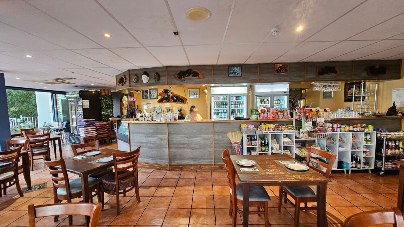 A cosy café interior with a tiled floor and a white panelled ceiling. In the foreground, wooden tables and chairs are set with placemats. The counter in the centre has a variety of items on display, with a person standing behind it. Shelves on the right hold various packaged goods. The left side features large windows with a view of greenery outside. The walls are adorned with decorative masks and framed pictures.