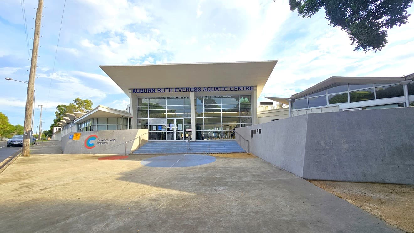 Concreted open space in front of Auburn Ruth Everuss Aquatic Centre. The facility is a large white brick building with glass elements on the front wall. The concrete wall on the left with green, blue, and red color lines forming a C beside ‘CUMBERLAND COUNCIL’ and the right concrete wall with black bar tiles distinguishes the arrival area. In the middle are the concrete stairs with metal handrails that lead to the glass entrance wall with a blue ‘AUBURN RUTH EVERUSS AQUATIC CENTRE’ detailed.
