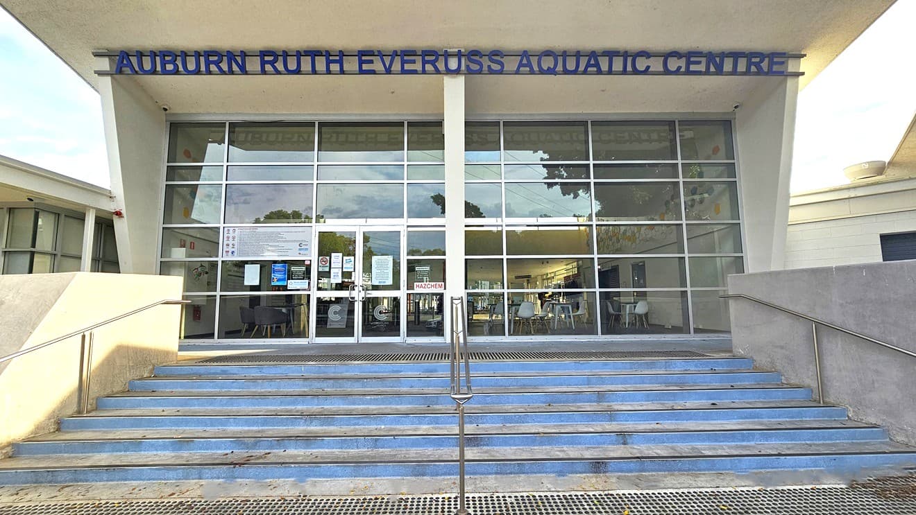 The stairs with metal handrails have light blue painted steps. At the bottom and top of the stairs are metal gratings for water drainage. The entrance door is on the left side, surrounded by varying kinds, colors, and sizes of informational signs and adverts. Two light gray lines forming a C are on the bottom of the glass entrance doors. On the top of the front wall is a blue ‘AUBURN RUTH EVERUSS AQUATIC CENTRE.’ Seen through the glass door and wall are the chairs, tables, and doors inside.