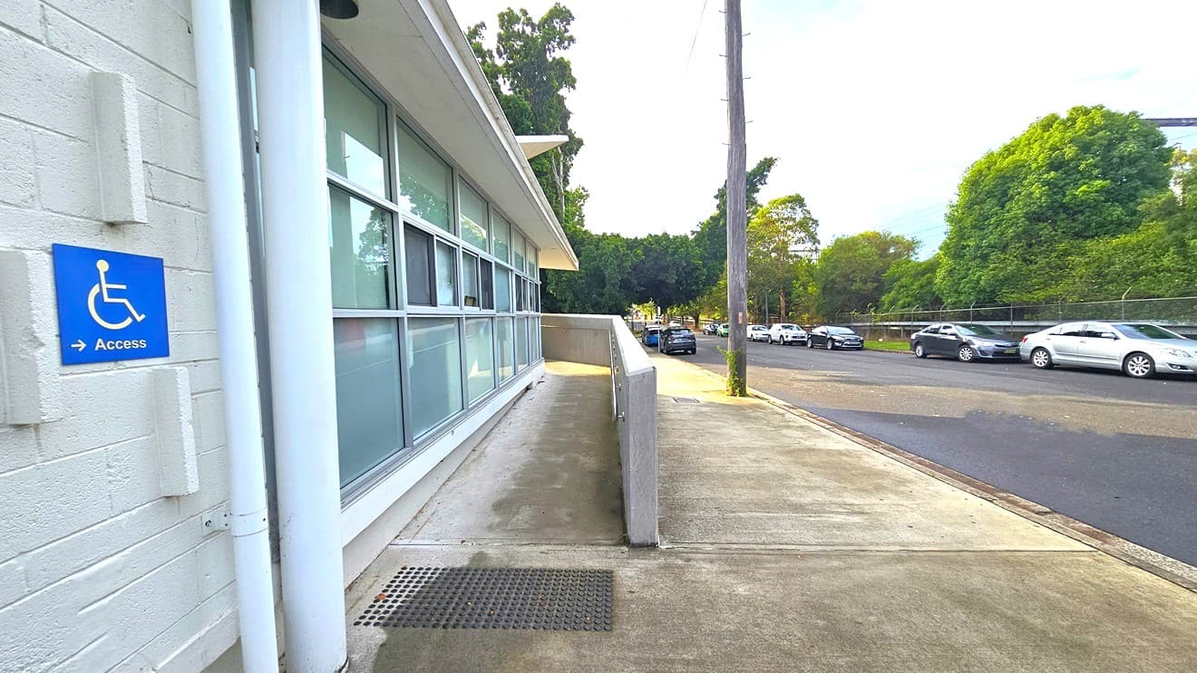 Ramp access marked by a blue square sign with a white image of a person in a wheelchair placed on the white brick wall. On the sides of the sign are three bricks protruding from the wall. Near it are small and large white pipes connected from the roof to the floor. Next to the pipes are blurry square glass windows with white metal frames. The ramp access has stainless steel handrails mounted on the right concrete wall. Several cars of different colors are parked on each side of the road.