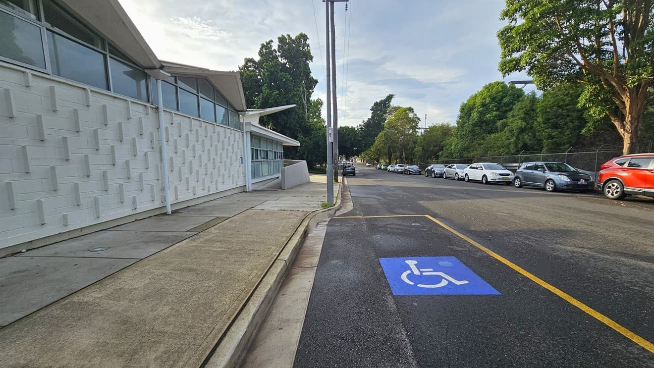 Further to the back of the left concrete wall with ramp access is the accessible car parking space for persons with mobility disabilities. The accessible car parking space has a blue image with a white wheelchair placed on the road and is separated by a yellow rectangular line. The white wall of the building has several protruding rectangular, narrow white bricks, while the upper part is made of glass windows with white metal frames.