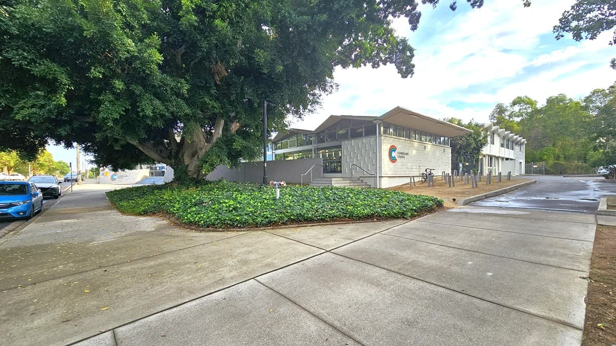 A garden filled with green plants, a large tree at the center, and a black lamp post features the right front area of the facility. There is a short staircase with metal handrails leading to another entry point to the centre. The left white brick wall has narrow rectangular white bricks protruding from the wall while the right brick wall has green, blue, and red color lines forming a C beside ‘CUMBERLAND COUNCIL.’ The space in front is filled with pairs of grey wood in an upright position.