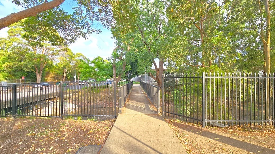Concrete pedestrian bridge from the right of Auburn Ruth Everrus Aquatic Centre with a black metal fence on each side. Under the bridge is a waterway that starts from the left and continues to the right and is marked by a higher black metal fence. Tall trees with abundant foliage offer significant shade to this walkway.