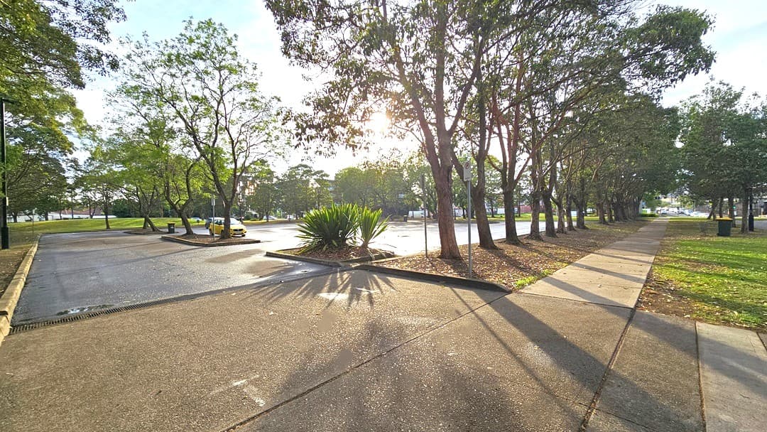 Extensive car park from the right side of the facility. Around the car park are trees lined on the edge of the garden. A No Parking sign with a red arrow at the bottom pointing towards the entry space to the extensive car park can be found on the right of the main entrance. Near it are two green plants from the palm family. Large grey trash bins with yellow covers are placed inside the car park and on the side of the concrete walkway.