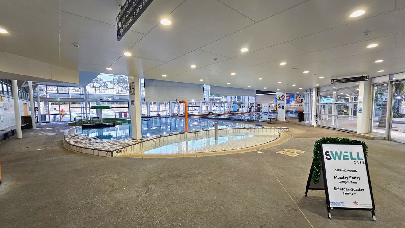 Leisure and Toddler Pool with a beach entry, an umbrella fountain, and stairs with handrails. To the right is a free-standing banner for the Cafe equipped with a Christmas garland. To the far right is another pool. The walls in the middle and the right are made of glass. Banners, safety warning signs, and directional signs are visible. The floor has concrete tiles, and the ceiling is all white with LED lights.