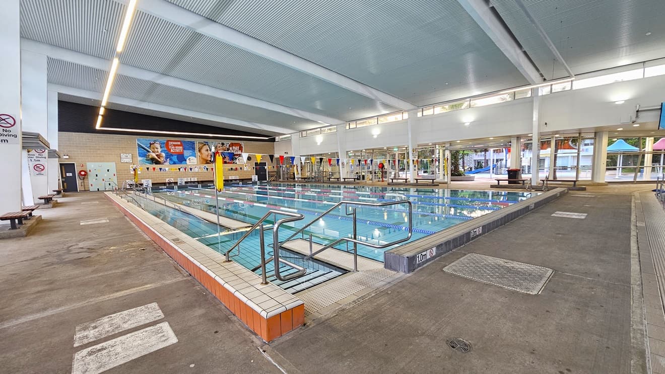 The 25m pool is equipped with steps and a ramp with handrails for entry. Truncated domes are installed before the steps. The pool is surrounded by a raised pool deck with markings denoting the pool depth. Floating ropes and colorful buntings run across the pool. Bench chairs are arranged on all sides of the pool. The middle wall features cream bricks, with large banners and security warnings affixed. The ceiling features a corrugated design with exposed metal beams and fluorescent lights.