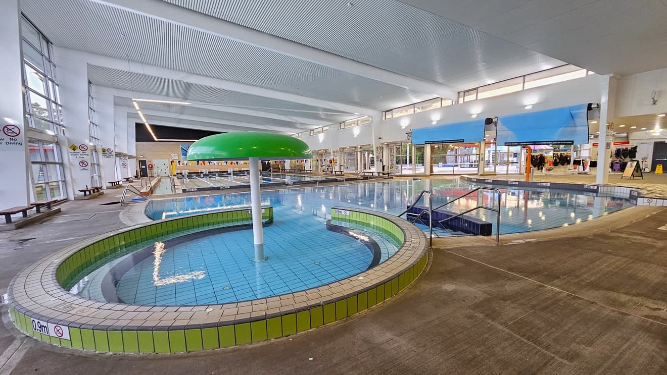 The Leisure and Toddler Pool has a stepped entry with handrails and a green umbrella fountain. To the right, banners and swimwear for sale are displayed near the pool deck. A rolled blue mat is positioned overhead. Adjacent is the 25m pool, equipped with a ramp and steps with handrails. Bench chairs are arranged on the left wall against the glass window. The ceiling features a white corrugated design with exposed metal beams and fluorescent lights.