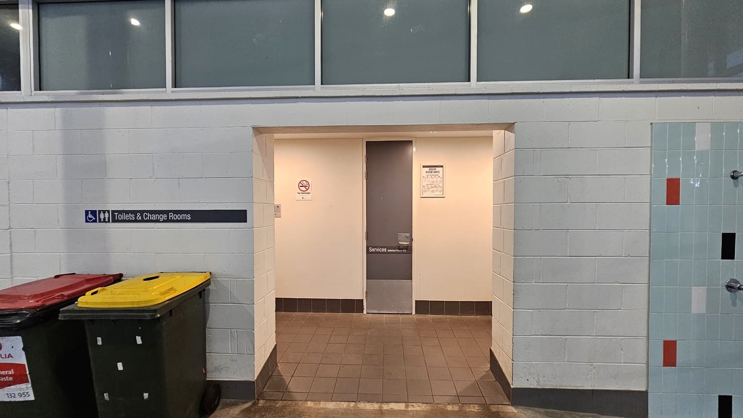 The accessible toilets and change rooms feature a white wall with a brick design. A signage is affixed to the left side of the wall. Two large rubbish bins are on the left. On the right is an outdoor shower area with multi-color brick tile accents. The upper wall features all glass, and the floor has brown tiles. Inside, the grey door is placed between two signages.