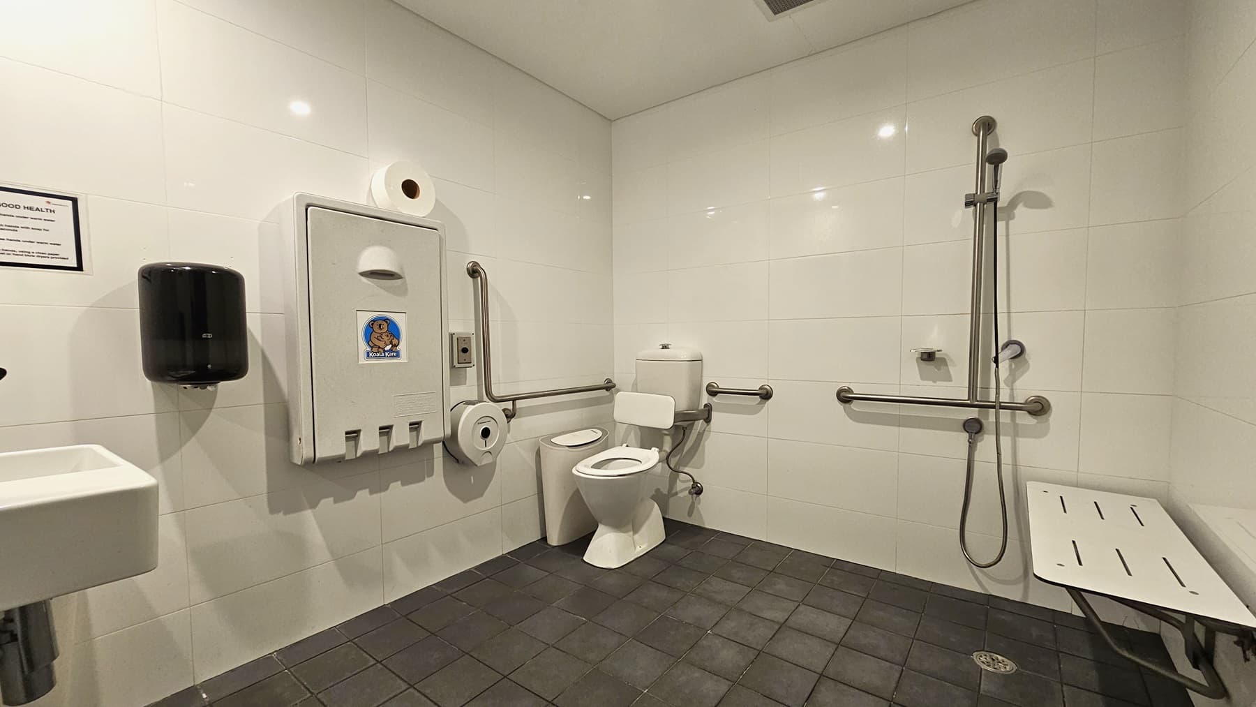 Inside the accessible bathroom, to the left is a toilet bowl equipped with hand grabs. A rubbish bin is placed beside it. A tissue dispenser, a foldable change table, and a liquid hand soap dispenser are mounted on the left wall. A wash basin and a poster are mounted on the wall on the left. To the right is a shower with handrails. A bench is positioned against the right wall. The floor has grey tiles, and the walls are white.