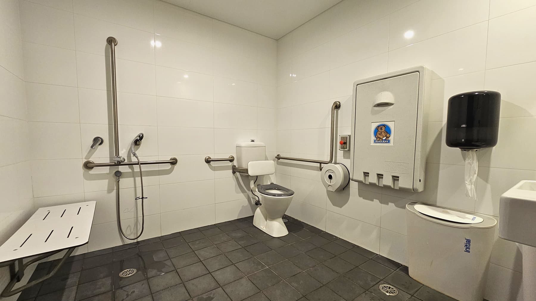 Inside the accessible bathroom, to the right is a toilet bowl equipped with hand grabs. A rubbish bin is placed beside it. A tissue dispenser, a foldable change table, and a liquid hand soap dispenser are mounted on the right wall. A wash basin and a poster are mounted on the wall on the right. To the left is a shower with handrails. A bench is positioned against the left wall. The floor has grey tiles, and the walls and ceiling are white.