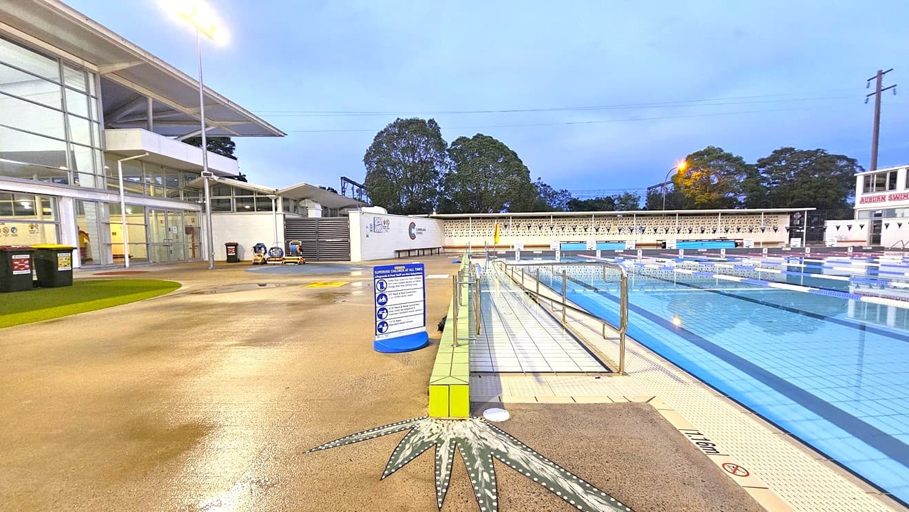 In the middle is the 50m 8-lane lap pool equipped with a ramp and step entry with handrails. Floating ropes and blue buntings run across the pool. Diving boards with covers are aligned on the raised pool deck on the right. Beyond the pool is an extensive tiered seating with shade. To the far left is a two-storey white building. Trees surround the area.