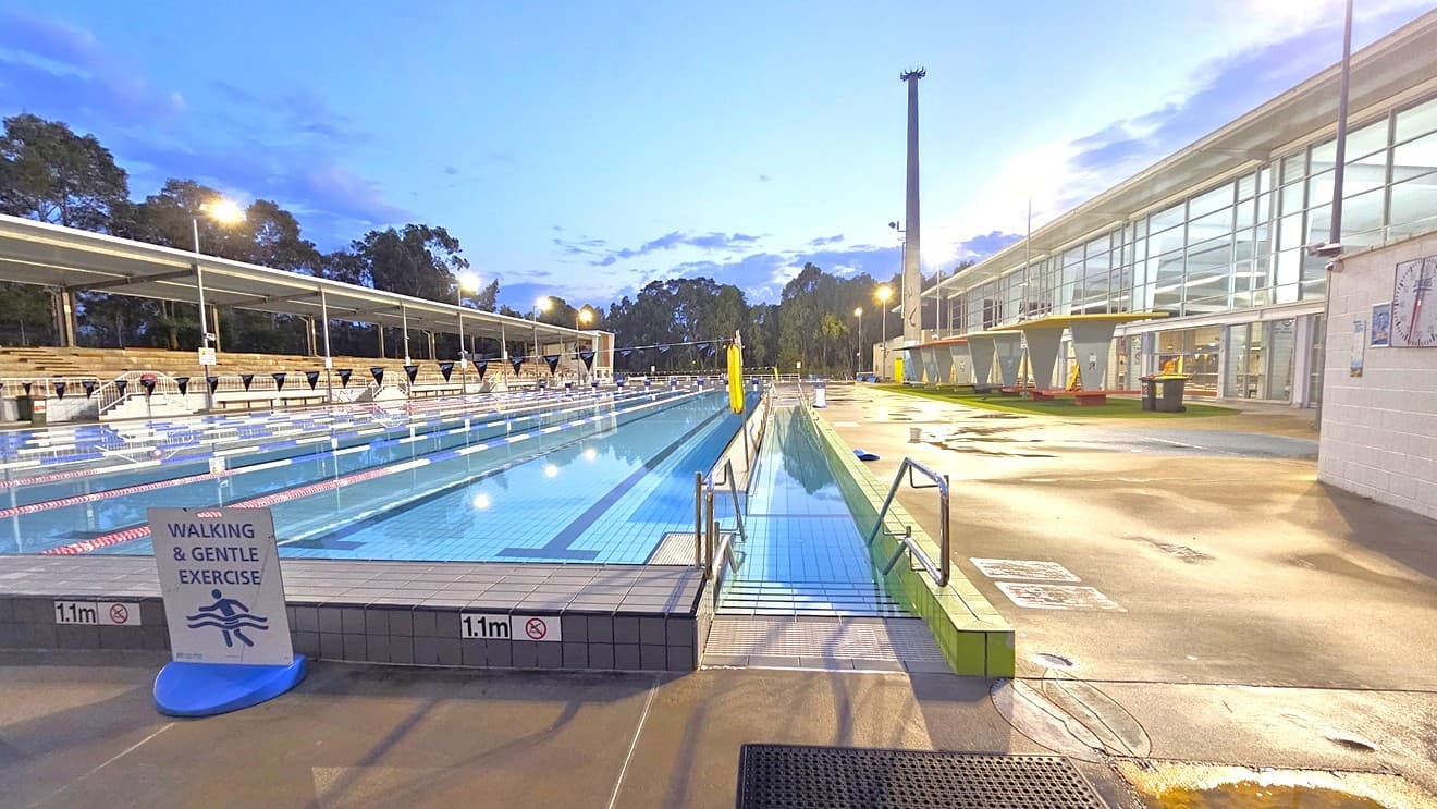 In the middle is the 50m 8-lane lap pool equipped with a ramp and handrails. A free standing banner stands on the pool deck. Floating ropes and blue buntings run across the pool. Truncated domes and a non-slippery mat adorn the ramp entrance. To the left is an extensive tiered seating with shade. To the right is the facility building with glass walls. Shade structures, seating, and rubbish bins are visible in the grass area.