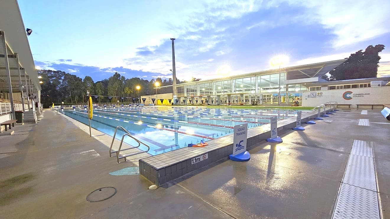 In the middle is the 50m 8-lane lap pool equipped with a ramp and handrails. A free standing banner stands on the pool deck. Floating ropes and blue buntings run across the pool. To the left is an extensive tiered seating with shade. To the right is the facility building with glass walls. Shade structures, seating, and rubbish bins are visible in the grass area.