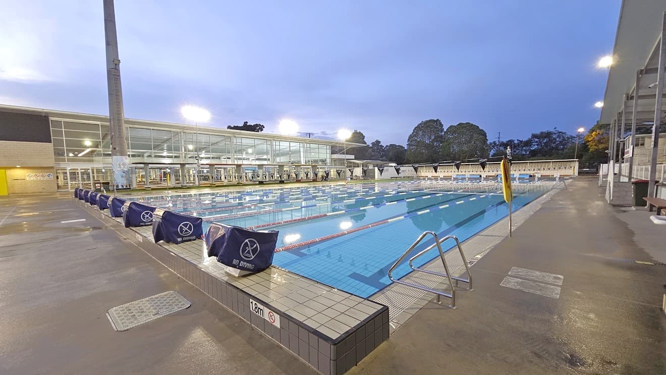 In the middle is the 50m 8-lane lap pool equipped with handrails. Diving boards with covers are arranged on the raised pool deck. A free standing banner stands on the pool deck. Floating ropes and blue buntings run across the pool. To the right is an extensive tiered seating with shade. To the left is the facility building with glass walls. Shade structures, seating, and rubbish bins are visible in the grass area.
