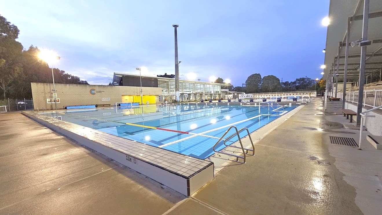In the middle is a dedicated polo sports pool equipped with handrails. In the middle is the goal set for the polo game. On the far end, diving boards with covers are arranged on the raised pool deck. Floating ropes run across the pool. To the right is an extensive tiered seating with shade. To the far left is the facility building with glass and brick walls. Tall floodlights are also visible.