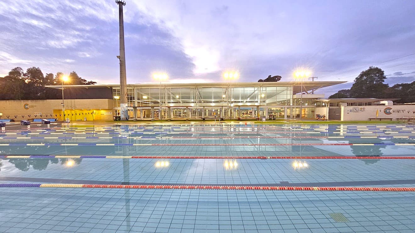 In the middle is the 50m 8-lane lap pool. Floating ropes and blue buntings run across the pool. Diving boards with covers are aligned on the raised pool deck on the left. Beyond the pool is the facility building featuring glass and brick walls. Shade structures are positioned in the manicured grass area in front of the building. Tall floodlights are positioned near the pool.