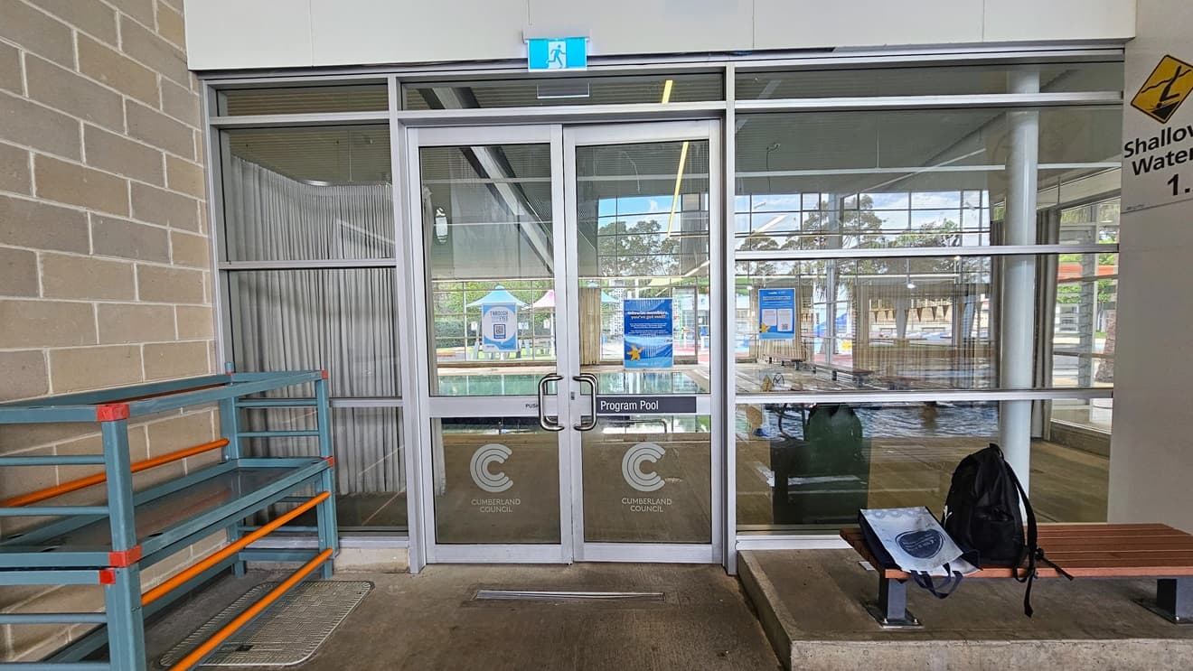 Entrance to the Program Pool is through an outward-opening glass door with hand grabs and facility stickers. A fire exit sign is mounted on top. To the left is a storage rack for belongings. To the right is a bench chair, positioned on a slightly elevated concrete platform against the glass wall. A backpack is placed on top. The upper right wall shows a "Shallow water" sign, and the left wall has brown bricks.