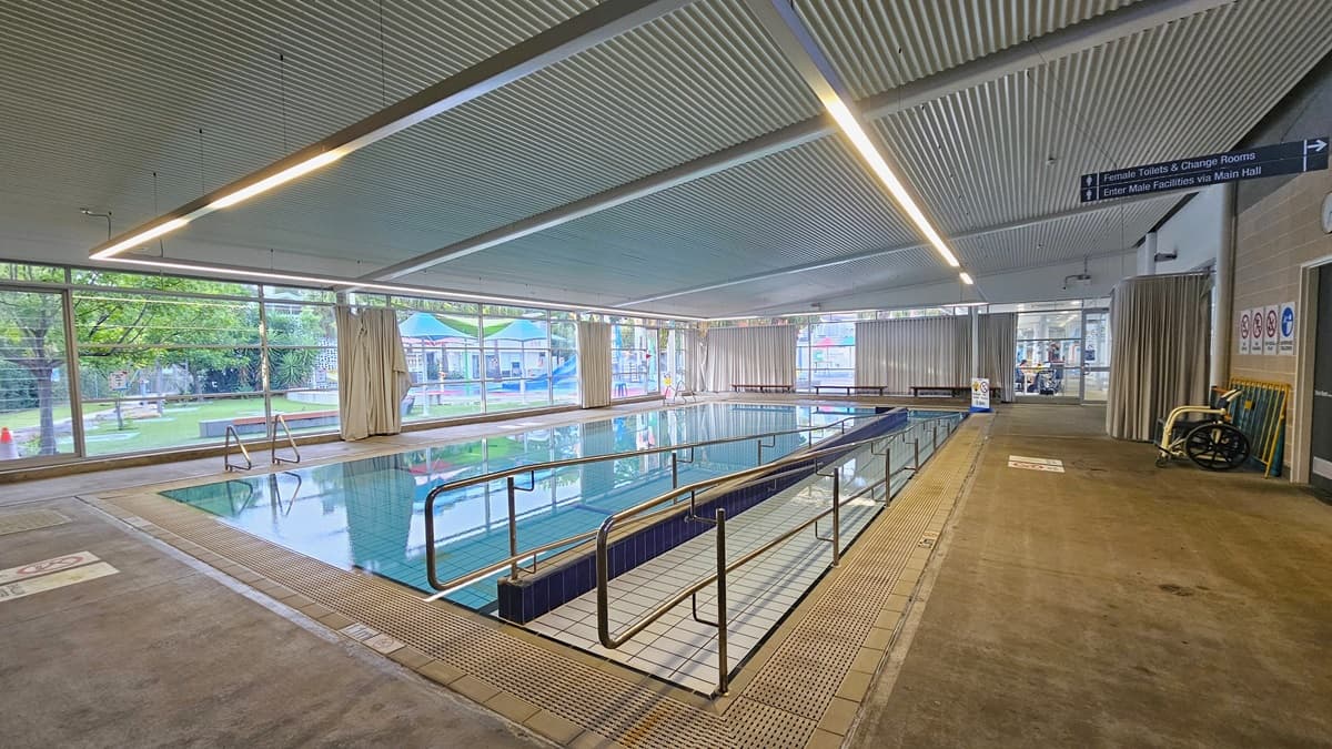 The Program Pool has a ramp entry with handrails on the right. A freestanding banner is positioned in the corner, and overflow grids line the pool. The left wall displays brown bricks and posters, while the right wall features glass with curtains. An outward-opening glass door leads to the Splash Pool. An aquatic wheelchair is placed near the brick wall on the right. The low-level corrugated ceiling is equipped with fluorescent lights, forming a continuous strip above the pool's perimeter.