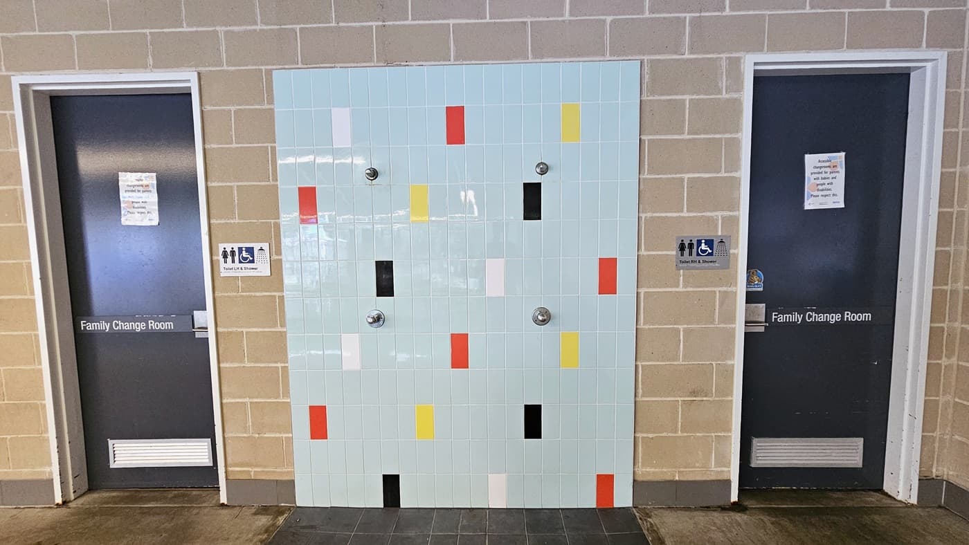 In the center of a brown brick wall is an outdoor shower with a wall adorned with multi-color tiles. Two grey doors, each marked with a "Family Change Room" sign, are present. Additional signage indicating accessibility, showers, and all-gender access is also mounted on the walls.