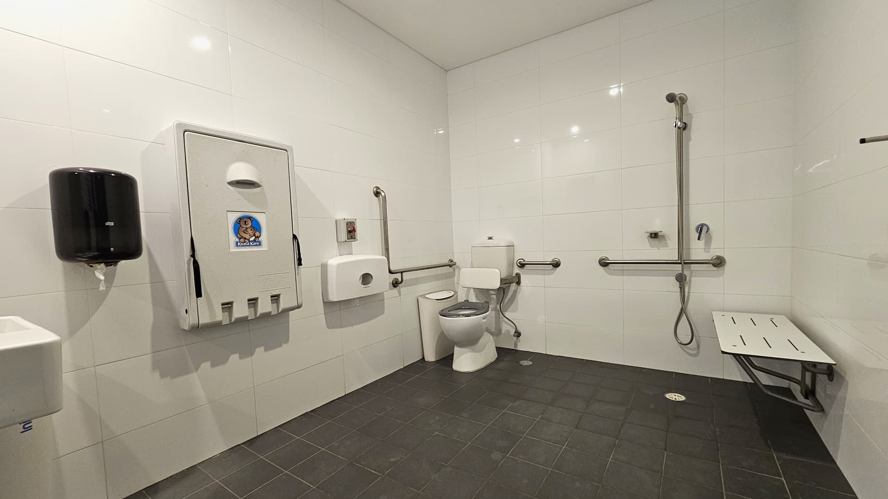 Inside the accessible change room, on the left is a toilet bowl equipped with hand grabs, accompanied by a rubbish bin. A tissue dispenser, a foldable change table, and a liquid hand soap dispenser are affixed to the left wall. A washbasin and a poster are mounted on the wall. To the right is a shower with handrails, and a bench is positioned against the wall. The floor is covered with grey tiles, and the walls and ceiling are white.