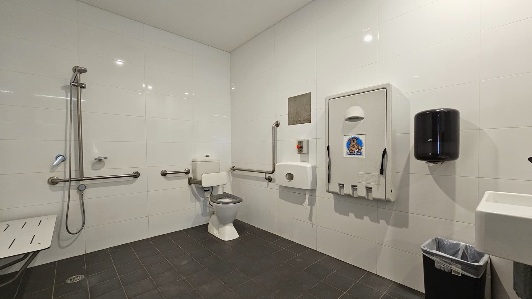 Inside the accessible change room, on the right is a toilet bowl equipped with hand grabs, accompanied by a rubbish bin. A tissue dispenser, a foldable change table, and a liquid hand soap dispenser are affixed to the right wall. A washbasin and a poster are mounted on the wall. To the left is a shower with handrails, and a bench is positioned against the wall. The floor is covered with grey tiles, and the walls and ceiling are white.