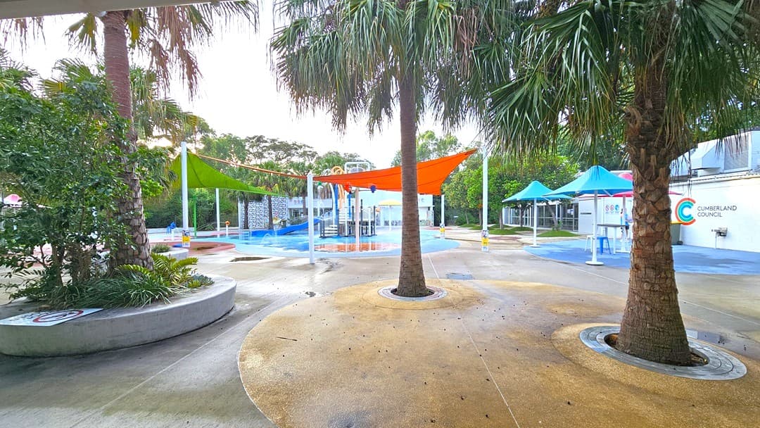 The outdoor Splash Park features palm trees and bright orange and green shade cloths. In the center is a blue slide with tipping buckets, water sprays, geysers, waterfalls, and fountains. Plastic tables and chairs are arranged under large blue umbrella shades on the right. The floor offers different shapes and colors. On the left, plants and trees are inside a round concrete planter box along with a safety sign. Further ahead, trees are visible.