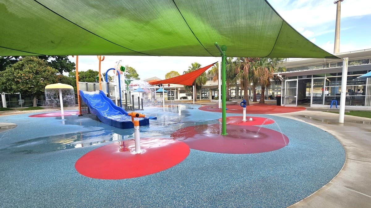 The Splash Park includes a blue slide, tipping buckets, water sprays, geysers, waterfalls, and fountains. Green and orange shade cloths offer shading. Tall palm and other trees border the left and right edges. The ground displays diverse shapes and colors. In the distance, a white building with a glass wall is visible on the right.