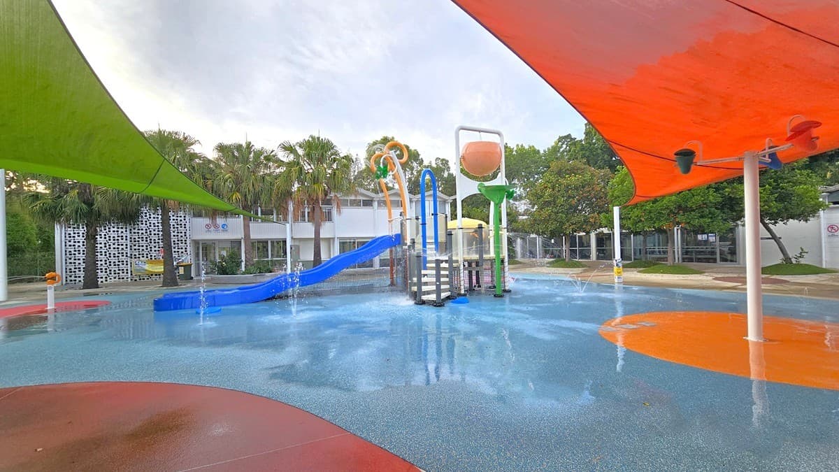 The Splash Park includes a blue slide, tipping buckets, water sprays, geysers, waterfalls, and fountains. Green and orange shade cloths provide shading. Tall palms and other trees border the left edges. The ground exhibits diverse shapes and colors. In the background, there's a white building with a glass wall. To the right, trees and neatly trimmed grass are visible.