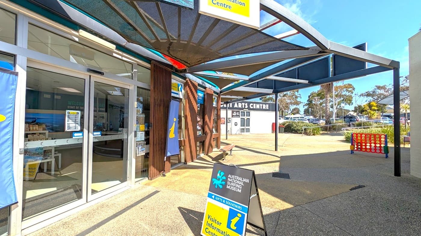 Entrance to the Australian National Surfing Museum on the left, with glass doors and informational banners. A signboard in front reads "Visitor Information Centre." A shaded area with a modern canopy extends overhead. In the background, the Surf City Arts Centre is visible, along with parked cars and trees. The ground is paved, and a large Connect Four game is set up on the right.