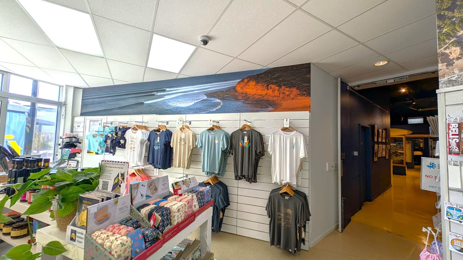 A shop interior with a display of T-shirts hanging on a white slatted wall in the centre. Above the shirts is a large mural of a coastal scene with waves and cliffs. On the left, there are shelves with various products, including jars and a potted plant. The ceiling is made of white tiles with fluorescent lights. To the right, there is a dark hallway leading further into the store. The floor is light-coloured.