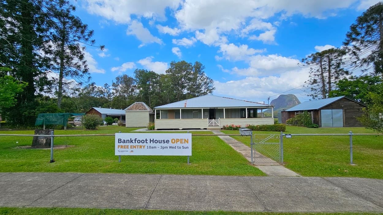 A single-storey house with a corrugated metal roof is in the centre, surrounded by a well-maintained lawn. To the left, there is a small wooden structure and a large tree. On the right, another wooden building with a metal roof is visible. A sign in the foreground reads "Bankfoot House OPEN" with visiting hours. A concrete path leads to the house, and a metal gate is part of the fence enclosing the property. Trees and a mountain are in the background.